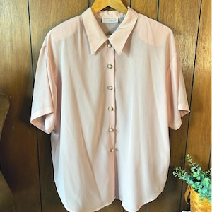 May include: A light pink button-down shirt with short sleeves. The shirt has a classic collar and is fastened with six pearlescent buttons. The brand name "impressions" is visible on the collar.
