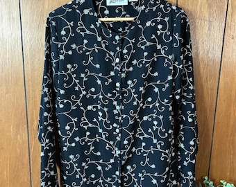 May include: Black button-down blouse with a delicate floral vine pattern in a light beige color. The long-sleeved shirt has a round collar and is hanging on a wooden hanger. The blouse is made of a soft, flowing fabric.