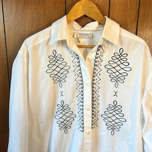 May include: White button-down shirt with long sleeves, hanging on a wooden hanger. The shirt features intricate black embroidered designs on the front and along the button line. The label reads "expressions sport".