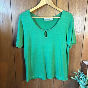 May include: A short-sleeved, bright green top with a keyhole detail at the neckline. The top is hanging on a wooden hanger. The fabric appears to be a knit material. A small tag is visible near the neckline.