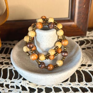 May include: Two beaded bracelets with wooden beads in shades of brown and cream, accented with small gold-colored beads. The bracelets are displayed on a small, off-white, ceramic dish. The bracelets are a jewelry item.