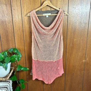 May include: A sleeveless top with a cowl neck, in shades of peach and coral. The top is made of a lightweight, open-knit fabric. It hangs on a wooden hanger against a wood-paneled wall. The top has a brown neckline.