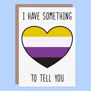 May include: A greeting card with a brown border and a white interior. The card features a heart-shaped design in yellow, white, purple, and black, representing the non-binary flag. The text "I HAVE SOMETHING TO TELL YOU" is printed above and below the heart.