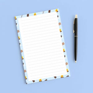 May include: A lined notepad with a light blue border featuring camping-themed illustrations, including marshmallows, campfires, and chocolate bars. A black pen with gold accents rests beside the notepad on a light blue surface.