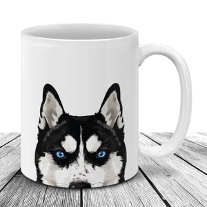 May include: White ceramic mug featuring a detailed illustration of a Husky dog's face. The dog has striking blue eyes, black and white fur, and upright ears. The mug has a curved handle and is set against a white background.