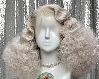 May include: A white wig with large, curly, voluminous waves. The wig is styled in a vintage, glamorous look. The wig is on a white mannequin head with a rhinestone necklace. The mannequin is on a silver sequined surface.