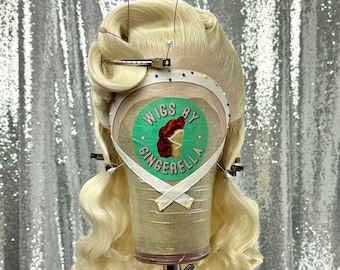 May include: A blonde wig with large, loose curls styled in a vintage updo. The wig is on a mannequin head and has a label that reads "Wigs by Cinderalla".