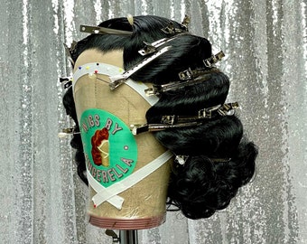Pode incluir: Uma peruca preta com risca lateral está penteada em um penteado vintage e presa com grampos prateados. A peruca está em uma cabeça de manequim com uma etiqueta verde e branca que diz "Wigs by Cinderella".
