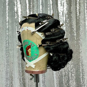May include: A black wig with a side part is styled in a vintage updo and secured with silver clips. The wig is on a mannequin head with a green and white label that says "Wigs by Cinderella".