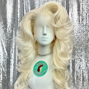 May include: A long, blonde wig with large, voluminous curls. The wig is styled with a high bouffant and falls in soft waves. The wig is on a mannequin head with a white background. The wig is labeled "Wigs by Gingerella."