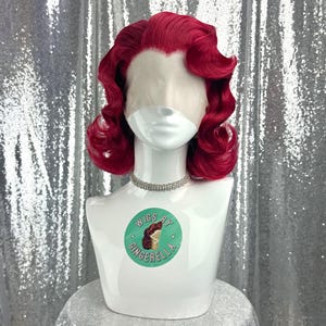 May include: A vibrant red wig styled with vintage curls, displayed on a white mannequin head. The mannequin wears a silver rhinestone choker. The base of the mannequin has a sticker that says "Wigs by Gingerella". The background is a silver sequined fabric.