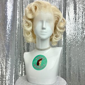 May include: A blonde wig with large curls styled on a white mannequin head. The wig is displayed on a white bust form with a diamante choker. A circular sticker on the bust reads "Wigs by Gingerella".