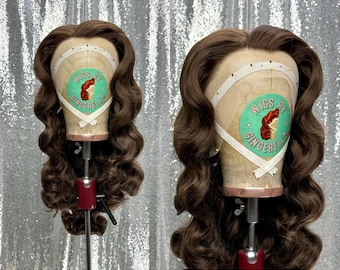 Peut inclure: Une perruque brune avec des boucles lâches coiffées en vague vintage. La perruque est sur une tête de mannequin avec un ruban de mesure blanc et une étiquette verte et blanche qui dit "Wigs by Cinderella".