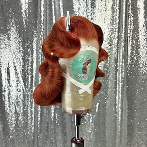 May include: A copper-colored wig styled with vintage waves, secured on a mannequin head. The wig is held in place with clips and pins. The mannequin head has a label that reads "Wigs by Gingerella". The background is a sparkly silver fabric.