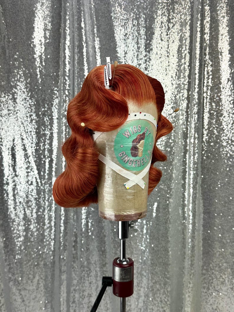 May include: A copper-colored wig styled with vintage waves, secured on a mannequin head. The wig is held in place with clips and pins. The mannequin head has a label that reads "Wigs by Gingerella". The background is a sparkly silver fabric.
