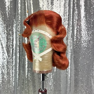 May include: A copper-colored wig styled with vintage waves, secured on a mannequin head. The wig is on a stand, and the text "KISS BY CINDERELLA" is visible on the head form. The background is a silver sequined fabric.