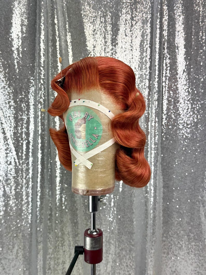 May include: A copper-colored wig styled with vintage waves, secured on a mannequin head. The wig is on a stand, and the text "KISS BY CINDERELLA" is visible on the head form. The background is a silver sequined fabric.