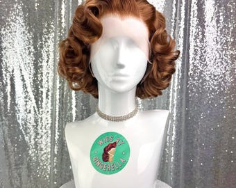 May include: A vintage-style auburn wig with curled ends is displayed on a white mannequin head. The wig is styled with a side part and soft waves. The mannequin wears a rhinestone choker and a sticker that says "Wigs by Gingerella". The background is a silver sequined fabric.