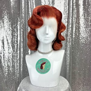 May include: A red-orange wig styled in vintage waves, displayed on a white mannequin head. The wig features a lace front and is accessorized with a silver rhinestone choker. The mannequin has a sticker that says "Wigs by Gingerella".