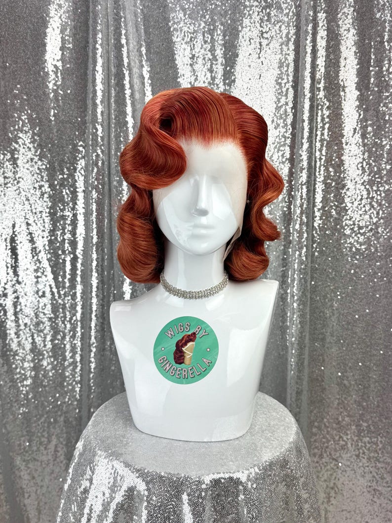 May include: A red-orange wig styled in vintage waves, displayed on a white mannequin head. The wig features a lace front and is accessorized with a silver rhinestone choker. The mannequin has a sticker that says "Wigs by Gingerella".