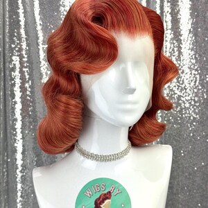 May include: A red-orange wig styled with vintage waves, displayed on a white mannequin head. The wig features a lace front and is accessorized with a silver rhinestone choker. A round sticker with the text "Wigs by Cinderella" is visible.