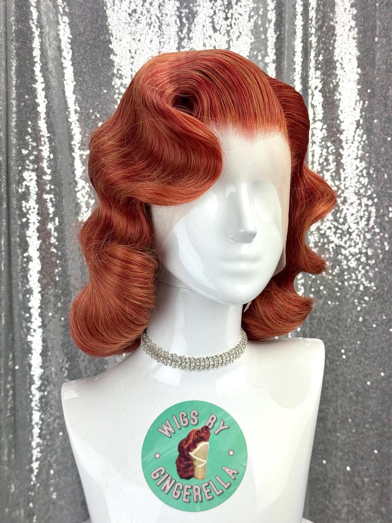 May include: A red-orange wig styled with vintage waves, displayed on a white mannequin head. The wig features a lace front and is accessorized with a silver rhinestone choker. A round sticker with the text "Wigs by Cinderella" is visible.