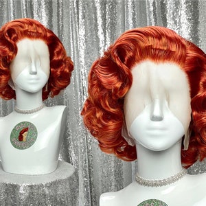 以下が含まれることがあります： 大きなカールのかかった波状の明るいオレンジ色のウィッグ。ウィッグは1950年代のヴィンテージスタイルでスタイリングされています。ウィッグは白いマネキンの頭に置かれています。