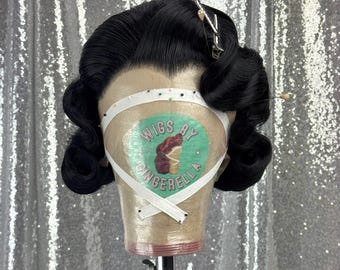 Pode incluir: Uma peruca preta com ondas vintage, fixada em uma cabeça de manequim. A cabeça é coberta com uma tira branca e o texto "Wigs by Gingerella" é visível. Grampos de cabelo prateados e um alfinete decorativo são colocados no cabelo.