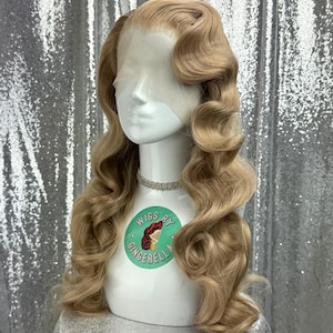 May include: A light blonde wig with long, voluminous curls styled in a vintage wave pattern. The wig is displayed on a white mannequin head with a delicate silver necklace. The mannequin has a sticker that says "Wigs by Gingerella".