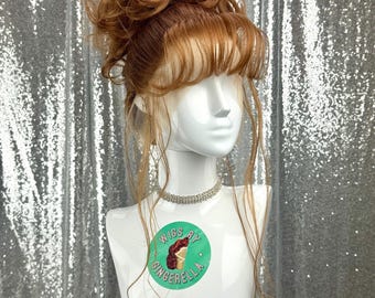 May include: A light brown wig with bangs and an updo, displayed on a white mannequin head. The wig has a natural-looking hairline and is styled with loose curls. A silver choker necklace is around the mannequin's neck. The mannequin has a green sticker that says "Wigs by Gingerella".