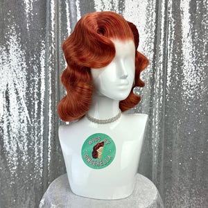 May include: A red-orange wig with vintage-style waves is displayed on a white mannequin head. The mannequin wears a silver rhinestone necklace. The base of the mannequin has a sticker that says "Wigs by Gingerella". The background is a silver sequined fabric.