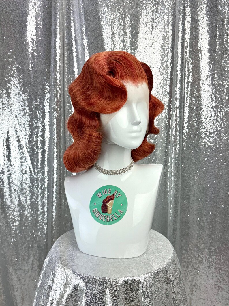 May include: A red-orange wig with vintage-style waves is displayed on a white mannequin head. The mannequin wears a silver rhinestone necklace. The base of the mannequin has a sticker that says "Wigs by Gingerella". The background is a silver sequined fabric.