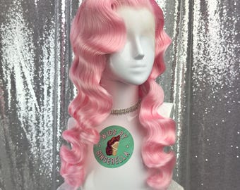 May include: A light pink wig with voluminous, wavy curls styled in a vintage fashion. The wig is displayed on a white mannequin head with a silver necklace. The mannequin has a green and white sticker that says "Wigs by Gingerella". The background is a silver sequined fabric.