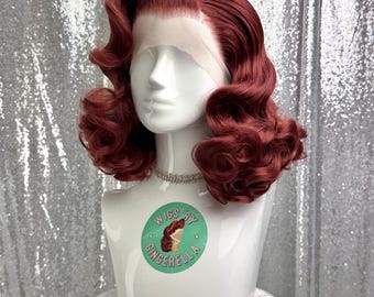 May include: A burgundy-colored wig styled with vintage waves, displayed on a white mannequin head. The wig features a lace front and is adorned with a small, sparkling necklace. The mannequin has a sticker that says "Wigs by Gingerella".