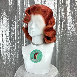 May include: A red-orange wig styled in vintage waves, displayed on a white mannequin head. The wig features a side part and a natural-looking hairline. The mannequin wears a silver choker and has a circular sticker that says "Wigs by Gingerella".