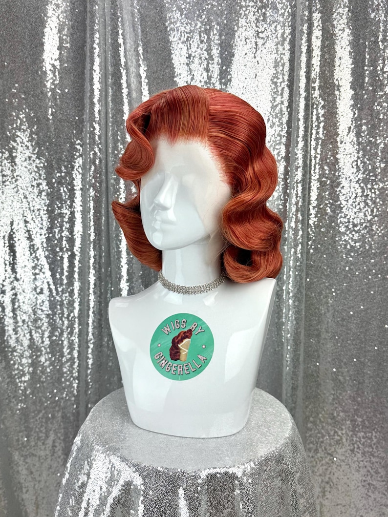 May include: A red-orange wig styled in vintage waves, displayed on a white mannequin head. The wig features a side part and a natural-looking hairline. The mannequin wears a silver choker and has a circular sticker that says "Wigs by Gingerella".