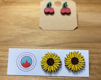 Peut inclure: Une paire de boucles d'oreilles en bois avec un motif de tournesol. Les boucles d'oreilles sont jaunes avec des centres bruns et sont présentées sur une carte blanche avec le texte "Candace's Craft Corner".