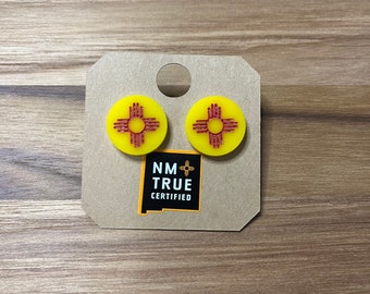 Peut inclure: Une paire de boucles d'oreilles jaunes et rouges représentant le symbole Zia, un symbole traditionnel du peuple Pueblo du Nouveau-Mexique. Les boucles d'oreilles sont sur une carte brune avec le texte "NM TRUE CERTIFIED".