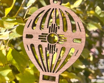 Peut inclure: Ornement en bois en forme de montgolfière avec un symbole Zia à l'intérieur. L'ornement est brun et a une ficelle attachée au sommet.