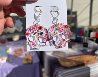 Peut inclure: Une paire de boucles d'oreilles en forme de crâne blanc avec un motif floral rose, rouge et violet. Les boucles d'oreilles ont des crochets argentés.