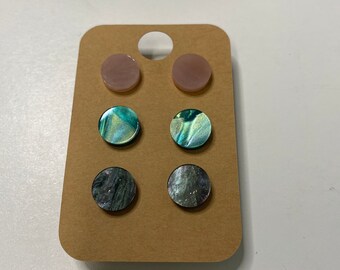 Peut inclure: Un ensemble de six boucles d'oreilles sur une carte marron. Les boucles d'oreilles sont rondes et faites de coquille irisée. Deux sont roses, deux sont vertes et deux sont noires.