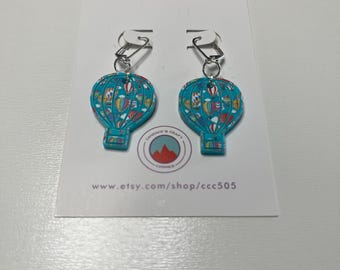 Peut inclure: Une paire de boucles d'oreilles montgolfières turquoise avec des détails colorés. Les boucles d'oreilles ont des crochets argentés et sont présentées sur une carte blanche avec le texte "Candace's Craft Corner" et une adresse de site web.