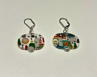 Peut inclure: Une paire de boucles d'oreilles en argent avec des breloques en acrylique transparent représentant un camping-car coloré. Les breloques sont remplies de diverses illustrations sur le thème du camping, notamment des arbres, une tente, un feu de camp et une guitare.
