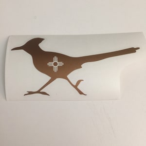 May include: A brown silhouette of a roadrunner with the symbol of the state of New Mexico on its chest. The symbol is white and features a sun with four rays.