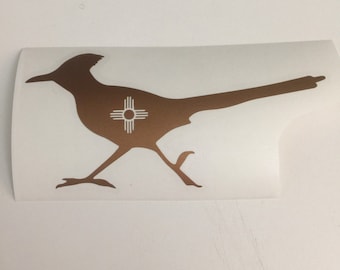Peut inclure: Une silhouette brune d'un coureur des routes avec le symbole de l'État du Nouveau-Mexique sur sa poitrine. Le symbole est blanc et représente un soleil avec quatre rayons.