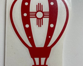 Peut inclure: Autocollant en vinyle rouge d'une montgolfière avec un symbole Zia stylisé à l'intérieur. Le ballon a des rayures blanches et des points rouges.