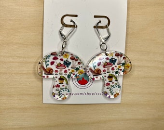 Peut inclure: Une paire de boucles d'oreilles en forme de champignon avec un fond blanc et un motif coloré de champignons, d'escargots et de feuilles. Les boucles d'oreilles sont en acrylique transparent et ont des crochets argentés.