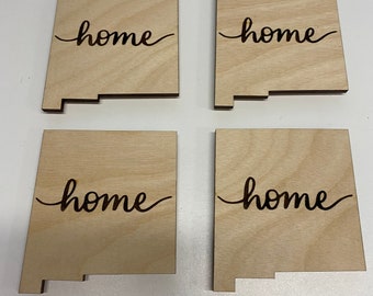 Peut inclure: Quatre sous-verres en bois en forme de l'état du Nouveau-Mexique avec le mot "home" écrit en cursive sur chaque sous-verre.