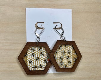 Peut inclure: Une paire de boucles d'oreilles hexagonales en bois avec un motif de nid d'abeilles et d'abeilles à l'intérieur. Les boucles d'oreilles sont brunes avec des crochets argentés.