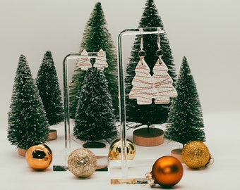 Peut inclure: Une paire de boucles d'oreilles en forme de sapin de Noël blanc avec des accents dorés suspendues à un support en acrylique transparent. Les boucles d'oreilles sont entourées de sapins de Noël artificiels miniatures et de décorations dorées et brunes.